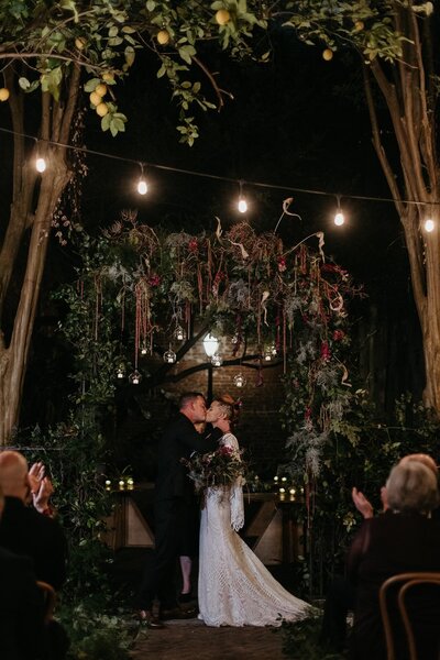 The newlywed couple kissing under the same floral arch, with guests clapping in the background.