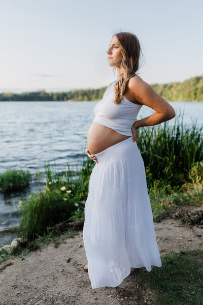 Pregnant woman standing by a serene lake in a flowing white dress, captured by South Bend maternity photographer.