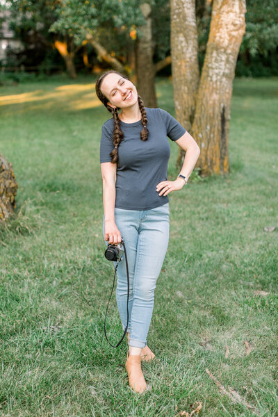 Girl in two braids, tshirt, and jeans with a film camera in her right hand and her left hand on her hip, smiling with her eyes closed