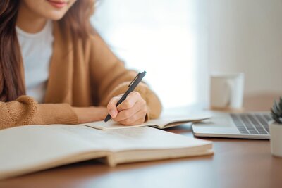 Woman in a brown sweater writing something in her journal at a desk with her laptop open 
