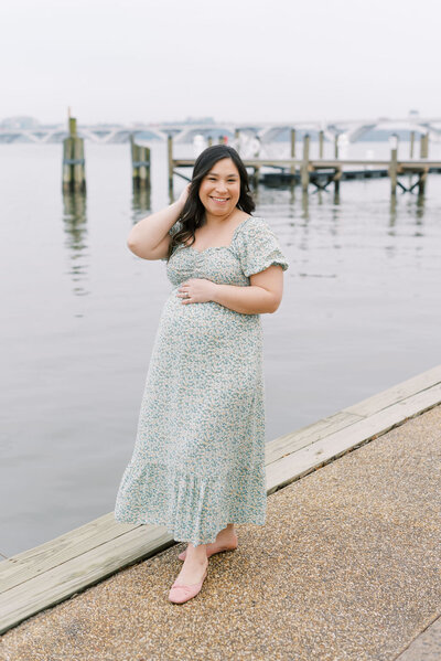 Richmond maternity photographer | Light and airy photo of a pregnant mom in a green floral dress playfully standing with her hand in her hair with the river behind her by Jacqueline Aimee Portraits