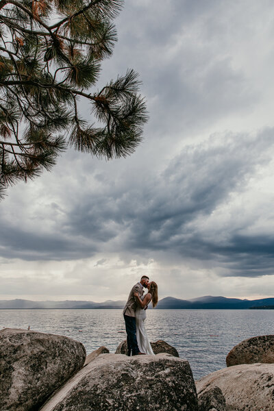 Couple sharing a kiss on the rocky shoreline at Sand Harbor in Lake Tahoe, with dramatic clouds and moody light over the lake and mountains.