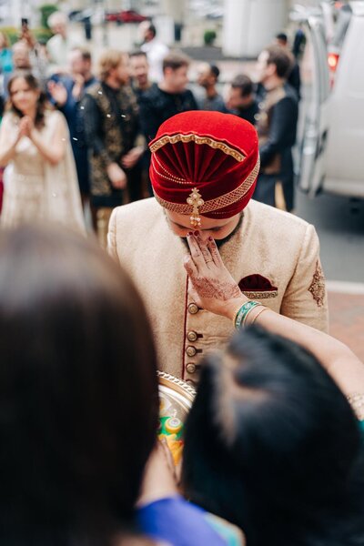A person in traditional indian wedding attire with a red turban covering their face with their hand while greeting guests.