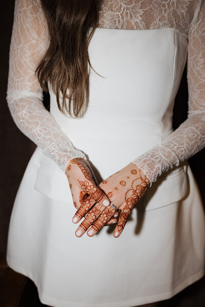 Brunette bride wearing a short wedding dress with lace details poses with her wedding ring and henna on hands.