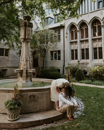 woman wearing white mid length dress standing by a old fountain leaning over and kissing man who is sitting below her