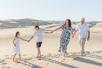 A mother, father, daughter, and son hold hands as they smile at each other while walking on the sand at Little Sahara recreation area.
