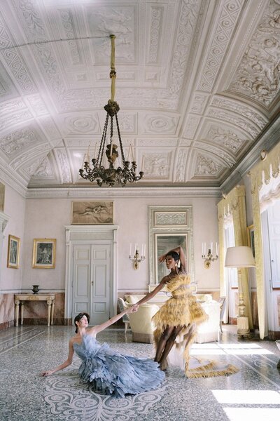 Two models in feathered Karen Sabag dresses perform gracefully in an elegant, ornate room with high ceilings and intricate moldings in villa Pizzo—an ideal setting for a luxury wedding photographer. A chandelier hangs from above, while sunlight streams through large windows, casting patterns on the polished floor.