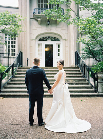 A bride and groom stand in front of the mansion at Burritt on the Mountain while the bride looks back at the photographer by Huntsville wedding photographer, Kelsey Dawn Photography