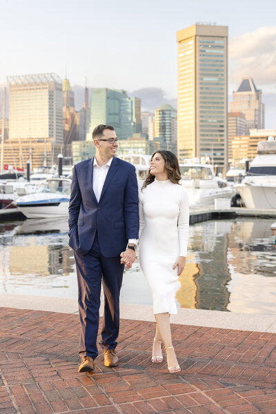 A couple holding hands walks along a waterfront, with boats and tall buildings in the background. The man is wearing a blue suit, and the woman is in a white dress. They are looking at each other, smiling.