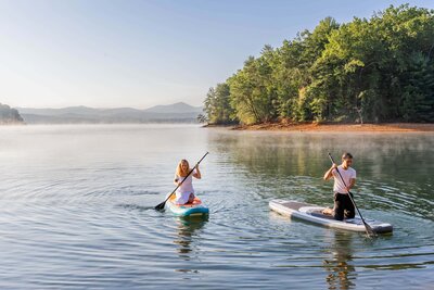 A bride and groom are kneeling on paddle boards paddling on a lake. They are wearing their wedding attire after eloping. There are lush green trees and a vast mountain range in the background.
