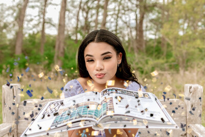 High school senior posing with open book and blowing confetti toward camera during senior session in Boerne, Texas.