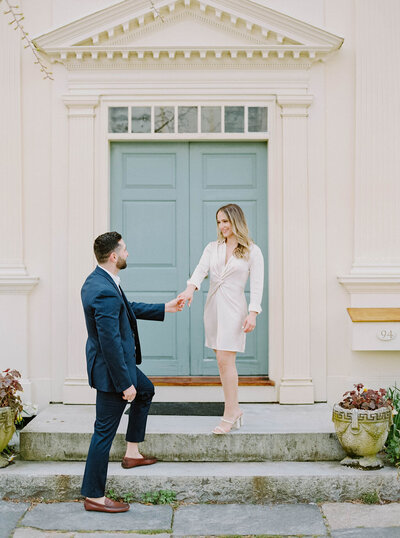 Rhode Island Wedding Photographer | A man in a dark suit helps a woman in a light dress step down from a stone porch. They are in front of a large teal door, with plants on either side.