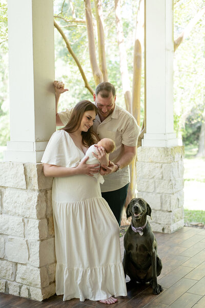 New parents hold their newborn on a cozy porch as their loyal dog sits nearby. A warm lifestyle newborn session by Moments in Light Photography.