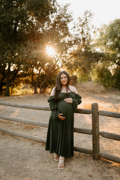 mom to be wearing green dress leaning against a wooden fence holding her belly during a sunset session at Horsethief Canyon Park in San Dimas, California 