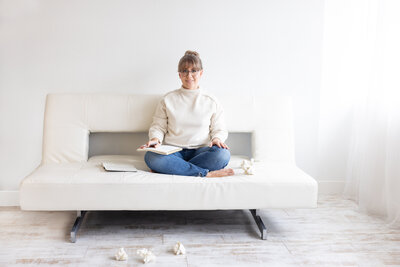 woman with her hair in a bun sitting cross-legged on a bed with a journal
