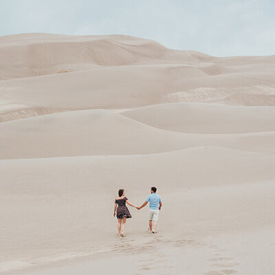 Couple walks holding hands across the landscape at Great Sand Dunes National Park in Colorado for their engagement photos