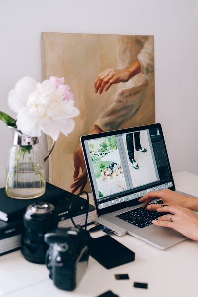 Hands typing on a laptop next to camera equipment, flowers, and a painting of hands