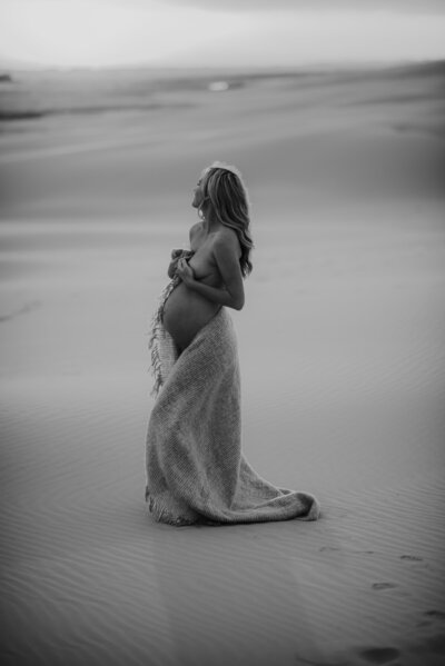 A woman stands on a sandy landscape, wrapped in a blanket, holding her pregnant belly. The image is in black and white.