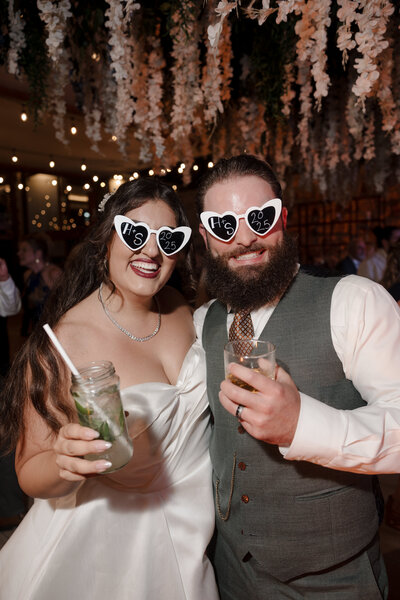 Hiba and her husband Shane on their wedding day in 2025. They're wearing white, heart-shaped sunglasses and holding drinks in their hands during their stunning outdoor wedding reception.