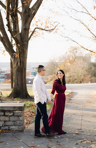 fall engagement photos with woman in a dark red dress holding hands with her fiance as they walk together on a path in richmond virginia wedding venues front lawn
