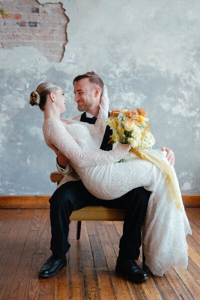 Woman in white gown sitting on mans lap. They are smiling at one another