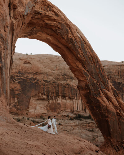 bride and groom walking under a large rock arch in the desert