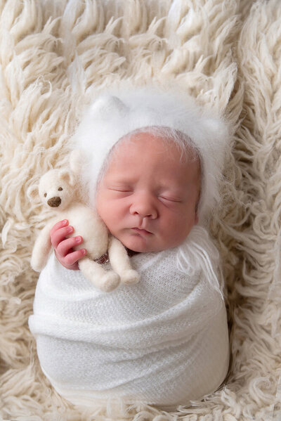 newborn baby boy wrapped in white wearing a white fuzzy bear hat, holding a tiny white bear for his newborn photography session.