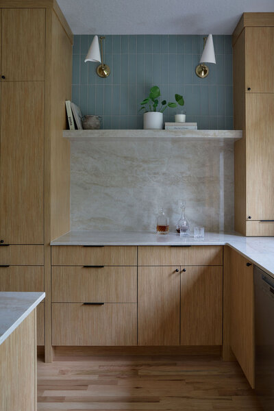 Kitchen with white oak cabinets, and slim simple black hardware. Taj mahal quartzite countertops that run up the backsplash to a quartz shelf with cookbooks and plants on it. Green subway tile backsplash above the shelf to the ceiling. Two sconces above the shelf that are modern, white and gold.