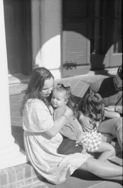 Black and white film photograph captured by a family photographer in Richmond, VA, showing a mother sitting on the front porch steps holding her toddler close in soft sunlight. Candid, emotional lifestyle moment.