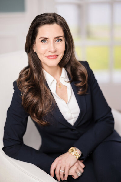 Executive headshot of a woman in a navy suit seated in a bright office environment, photographed for corporate branding.