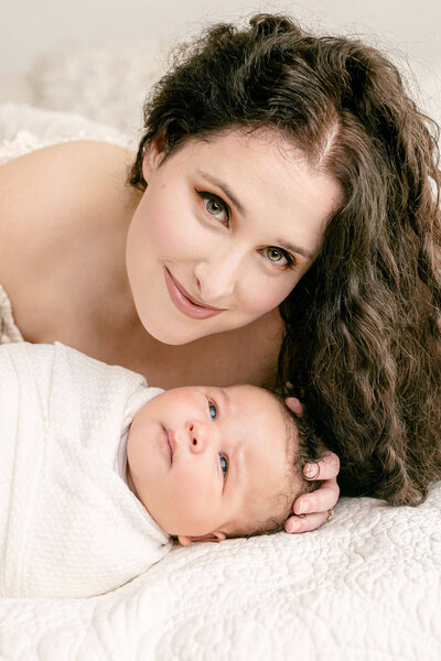 New Mom with long curly dark hair and wearing professional makeup. She is laying down on a bed with her face above her baby who is laying on the bed in front of her. She is looking at the camera with stunning green eyes and has a soft smile on her face. Baby is wrapped in white and his eyes are open and he is calm and looking up at his mom.
