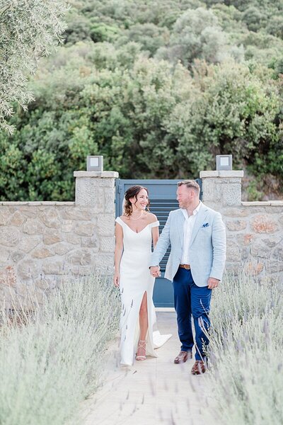 Bride and groom walk hand in hand through a romantic lavender-lined pathway at a private estate, dressed in modern destination wedding attire