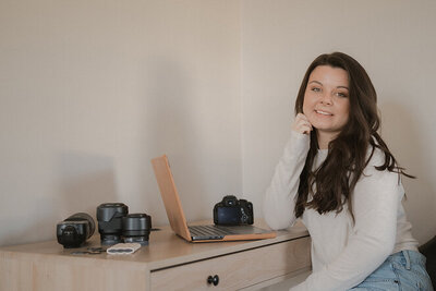 Michaela sitting at her desk with her laptop open, smiling at the camera. She is resting her chin on her hand.