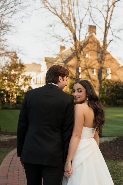 A couple walks hand in hand on a brick path in front of a historic estate as the bride looks back at the camera in a strapless gown, photographed for Grey Cove Designs wedding stationery clients.