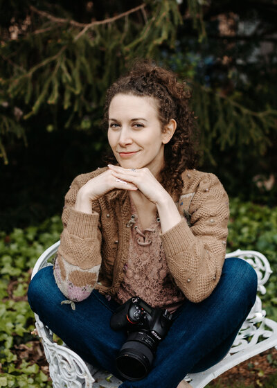 woman in brown sweater sits on white cast iron bench with nikon camera