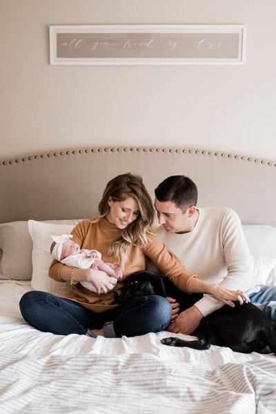 A Pittsburgh family photographer captures a tender moment of a couple sitting on a bed with a newborn and a black dog, showcasing the essence of family photography in Pittsburgh.
