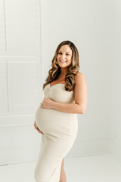 Smiling pregnant woman in beige tight dress that shows here growing belly.  The picture is taken in Lisa Levevre's studio in Atlanta.