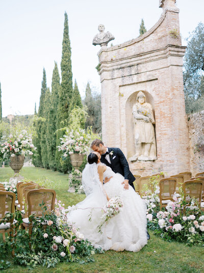 Villa Cetinale wedding in Tuscany Italy. Bride and groom kiss down aisle after getting married in gardens of Cetinale in Tuscay with ruffled dress, lush pink and greenery florals and Cetinale architecture