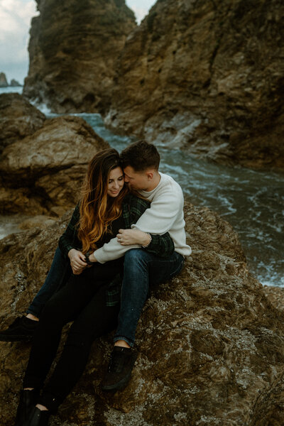 B sitting in between T's legs on a rock surrounded by the ocean, as T whispers into B's ear