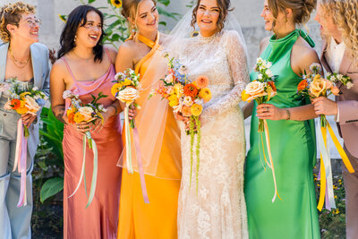 The bride stands smiling with her wedding party in front of the Ohio Statehouse in August 2024. She wears a long-sleeve lace gown and holds a colorful bouquet. Her wedding party are dressed in vibrant outfits—ranging from suits to dresses in pastel blue, pink, orange, green, and blush—and each holds a coordinating bouquet with ribbon streamers.