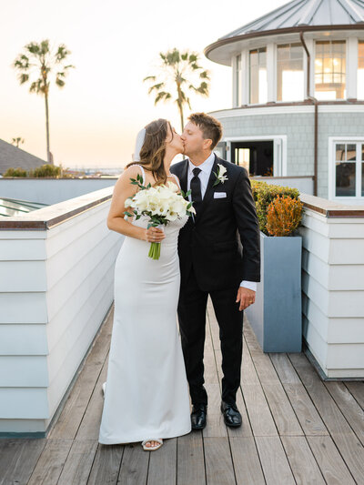 The bride and groom share a romantic kiss on the rooftop of Lido House in Newport Beach, surrounded by a golden sunset and ocean breeze with palm trees in the background