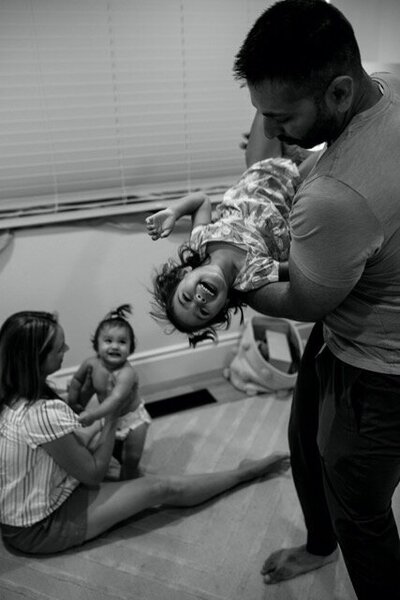 A dad swings his toddler daughter in his arms while she laughs and her baby sister looks on as mom is changing her. 