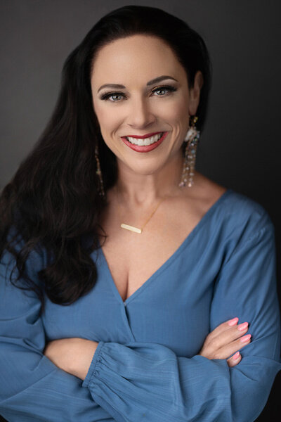 Smiling woman with long dark hair wearing a blue blouse and dangling earrings, arms crossed. She conveys confidence and warmth against a dark background.