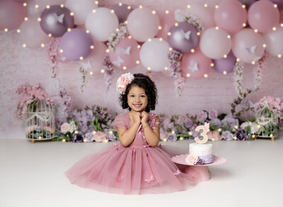 3rd birthday photo of girl in pink dress smiling at camera. Balloon backdrop, outfit and cake provided at Atlanta photo studio by Jenni Nikki.