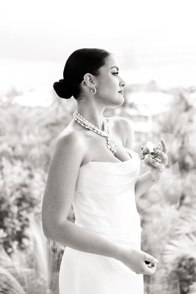 Black and white portrait of a bride in a strapless gown and statement pearls, applying perfume while overlooking tropical scenery in Riviera Maya, Mexico.