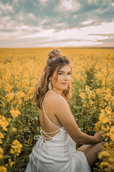 Senior girl with brown wavy hair sitting in a field of yellow flowers looking over her shoulder in a white dress with a soft smile