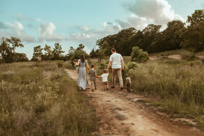 Mom in blue dress carries baby boy on hip while holding hands with her sons and husband with their dog as they walk off on dirt path during summer family session