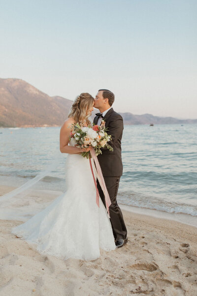 bride and groom standing on the beach while groom kisses bride's head