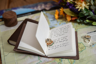 Open leather-bound vow books with handwritten wedding vows, gold wedding rings resting on the pages. The books sit atop a detailed map, with autumn florals in the background, symbolizing an adventure-filled Montana elopement.