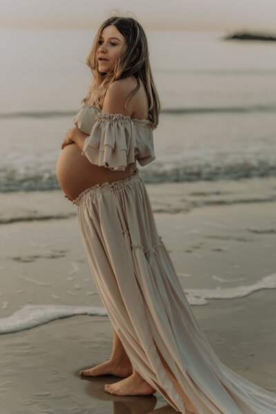Zwangere vrouw in jurk poseert aan het water op het strand tijdens fotoshoot